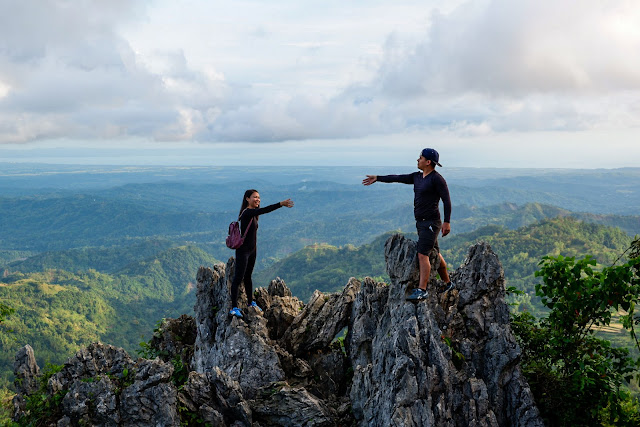 a man and a woman reaching out standing on top of the rocks in a mountainous place